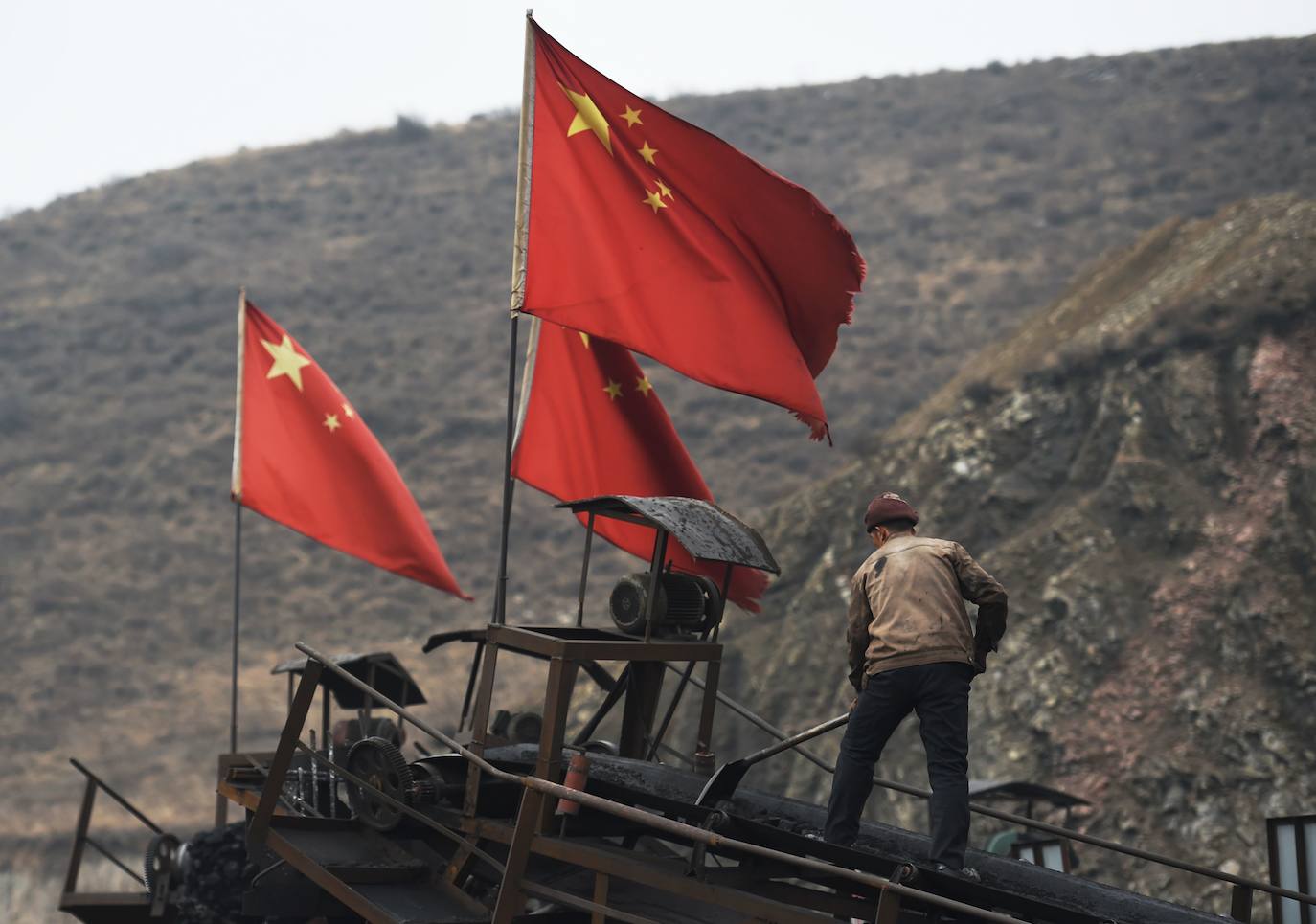 Esta foto de archivo tomada el 20 de noviembre de 2015 muestra banderas chinas junto a un trabajador que limpia una cinta transportadora utilizada para transportar carbón, cerca de una mina de carbón en Datong, en la provincia de Shanxi, en el norte de China. La promesa sorpresa de China de reducir su huella de carbono a cero para 2060 fue recibida con un aplauso cauteloso, pero un nuevo gasto en carbón para acelerar una economía golpeada por el virus amenaza con anular su audaz apuesta por llevar al mundo hacia un futuro con bajas emisiones de carbono.