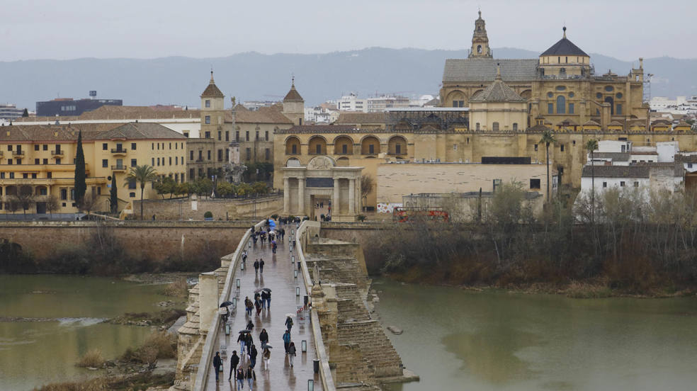 Varias personas se protegen de la lluvia con paraguas mientras pasean por el puente Romano de Córdoba con la Mezquita Catedral al fondo. 
