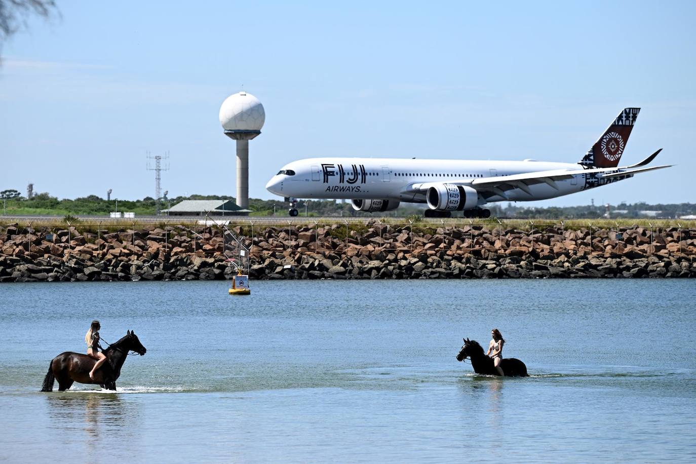 Dos mujeres a caballo se bañan en las aguas poco profundas de la bahía Botany con un avión de Fiji Airways en un aeropuerto del fondo, en Sydney, Nueva Gales del Sur (Australia). Las condiciones sofocantes continúan en esta zona del país con una ola de calor que va en aumento.