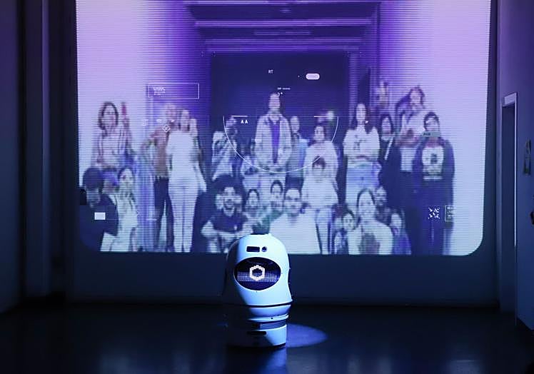 El robot junto a una pantalla donde se ven a todos los visitantes fotografiándolo.