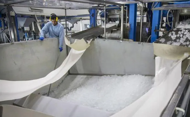 Imagen principal - Arrba, un operario vigila el proceso de producción en la planta. Abajo, a la izquierda, los bloques de hielo caen en la tolva tras ser cortados. A la derecha, envases de cubitos almacenados para su distribución.