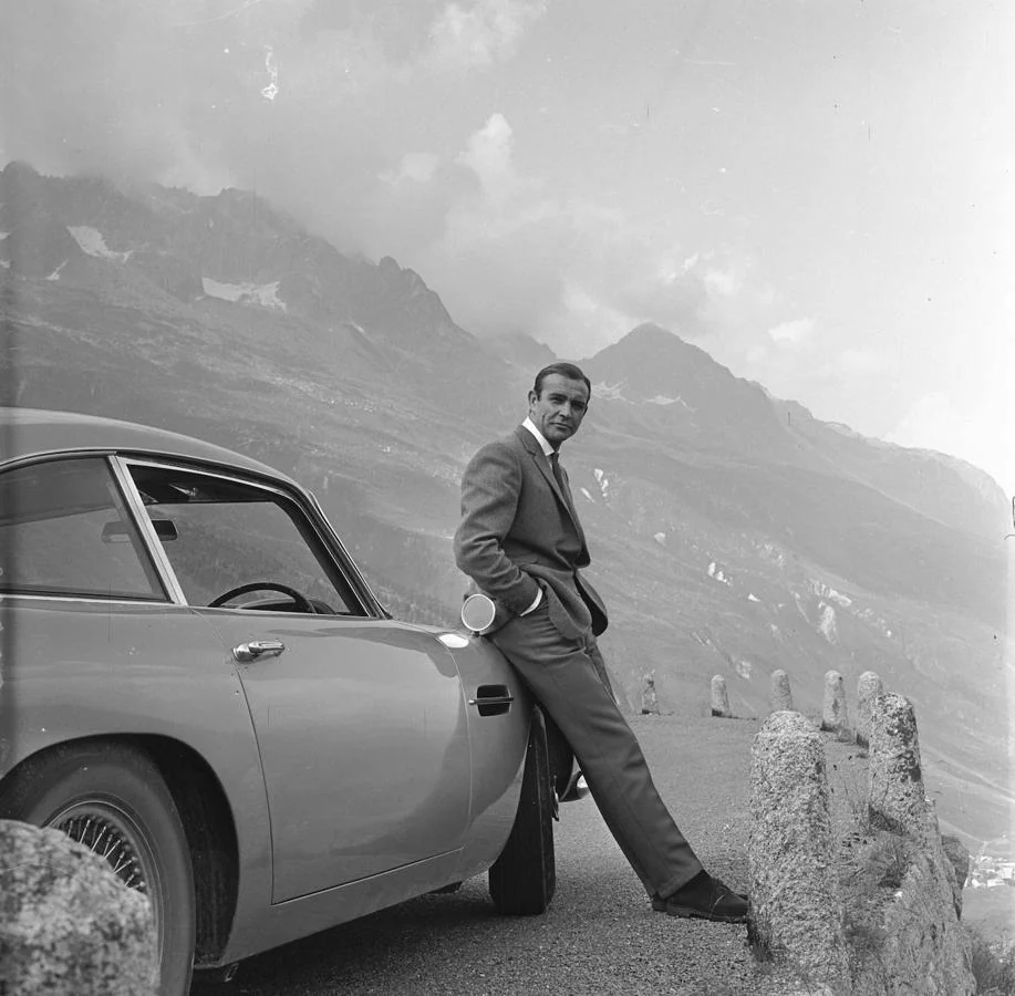 El actor Sean Connery descansando en el parachoques de su Aston Martin DB5 durante el rodaje de escenas para James Bond contra Goldfinger en los Alpes suizos.