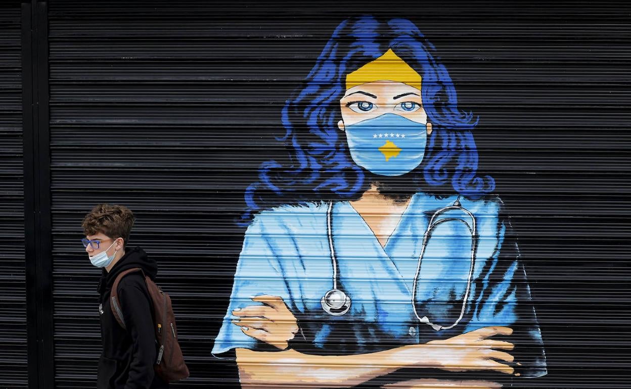 Un adolescente con la mascarilla medio bajada pasa por delante del grafiti de un sanitario