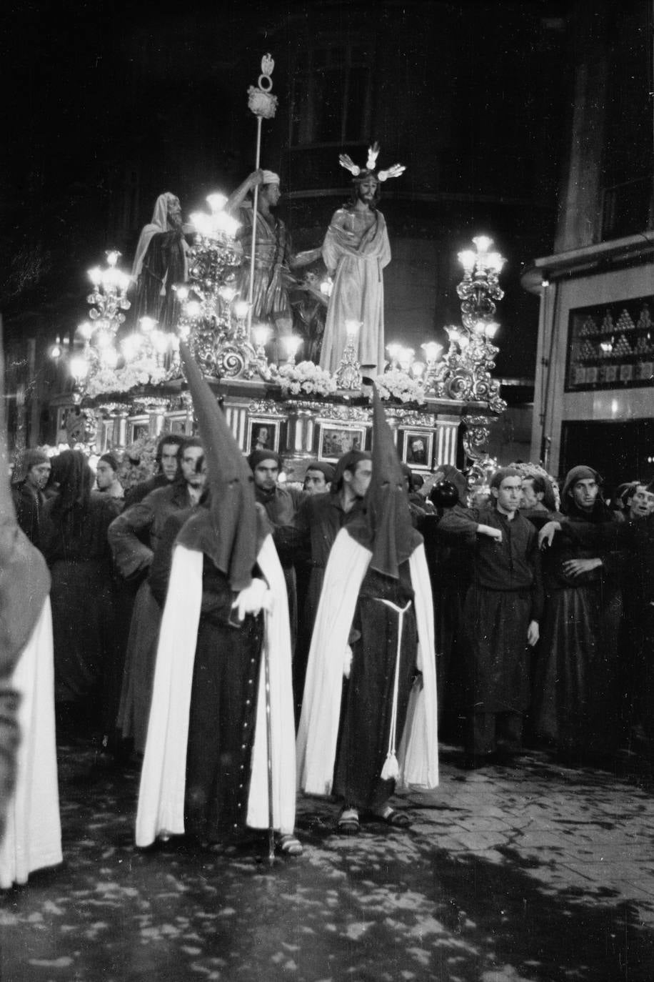 El trono de Jesús de la Sentencia aparece detenido en un momento de su salida procesional. Las imágenes todavía no han sido adaptadas para ser vestidas y figuran sobre el trono de corte clásico que construyó Nicolás Prados López en 1939 y que, en 1942, fue ampliado con nuevos arbotantes.
