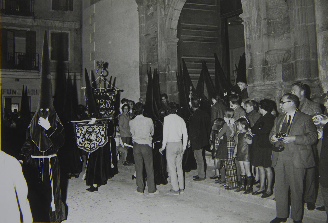 La Hermandad de las Penas, después de su estancia en la iglesia de San José, se trasladó al templo de San Julián, dado que el Obispado comunicó en los primeros meses de 1966 la venta de San José a una empresa constructora. En la fotografía, el cortejo de nazarenos de la sección del Cristo de la Agonía, ya con las túnicas de terciopelo que antecedieron a las actuales, saliendo desde San Julián el Martes Santo de 1969. Los tronos no salieron del interior de esta iglesia hasta 1992, por lo que lo hacían desde un tinglado.