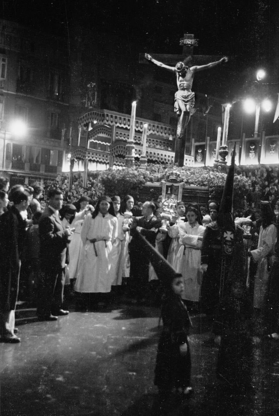 El trono del Cristo de Ánimas y Ciegos, detenido tras pasar ante la tribuna oficial de la época, que contaba con un palco cubierto por altos doseles, como se aprecia en la imagen. El trono es todavía provisional, aunque ya posee los cuatro hachones que todavía caracterizan al conjunto procesional de esta imagen, si bien durante varios años fueron sustituidos por faroles.