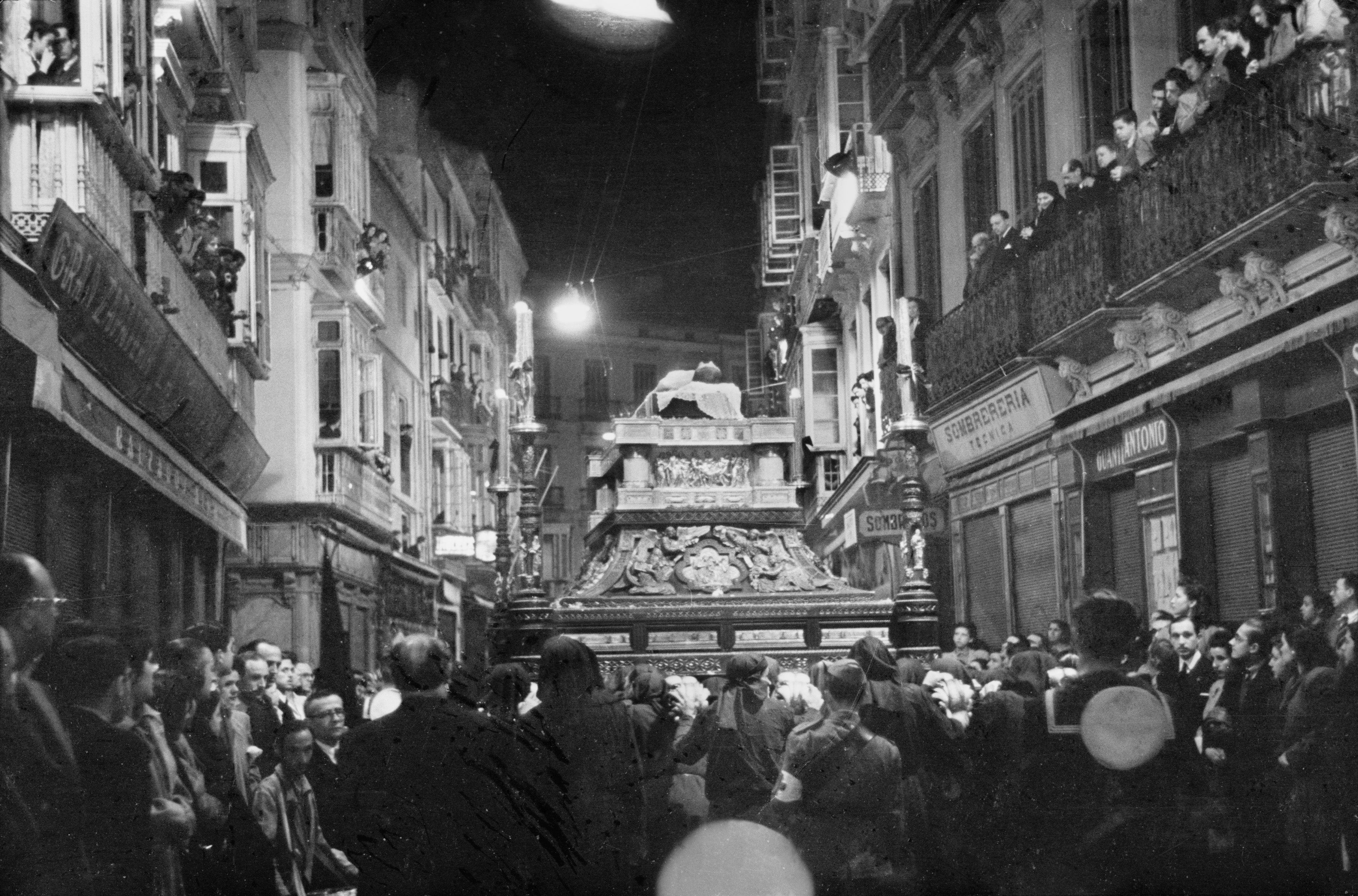 El trono del Santo Sepulcro discurre por la calle Granada después de efectuar el recorrido oficial. Era Viernes Santo de mediados de los años 40 del siglo pasado. Como puede observarse, en los ángulos ya figuraban los característicos hachones que se incluyeron al conjunto en 1943. Estas andas exhibían en su origen, en 1927, un largo fuste en cada una de las esquinas culminado con ocho candelabros.