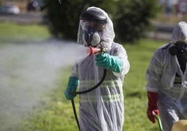 Dos operarios equipados con trajes protectores desinfectan una zona al aire libre.