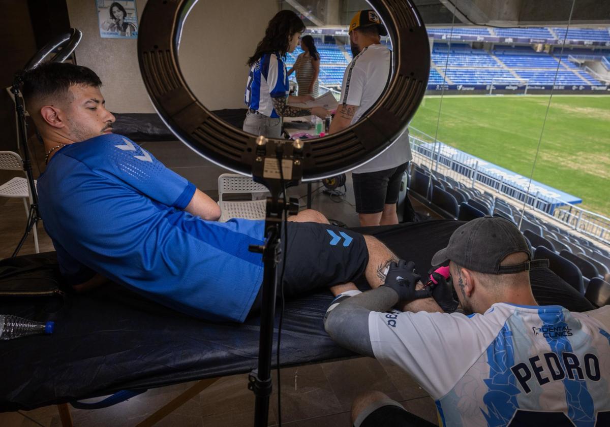 Imagen principal - Un aficionado del Málaga se tatúa la fecha del ascenso (22/06/24) debajo del escudo, a manos del tatuador Pedro. Dos aficionados muestran sus tatuajes. Detalle de uno de ellos.