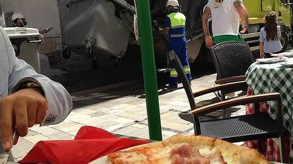 Unos comensales almuerzan una pizza en un restaurante del centro de la ciudad mientras que a escasos metros operarios de Limasa, provistos de mascarillas, recogen los contenedores de basuras.