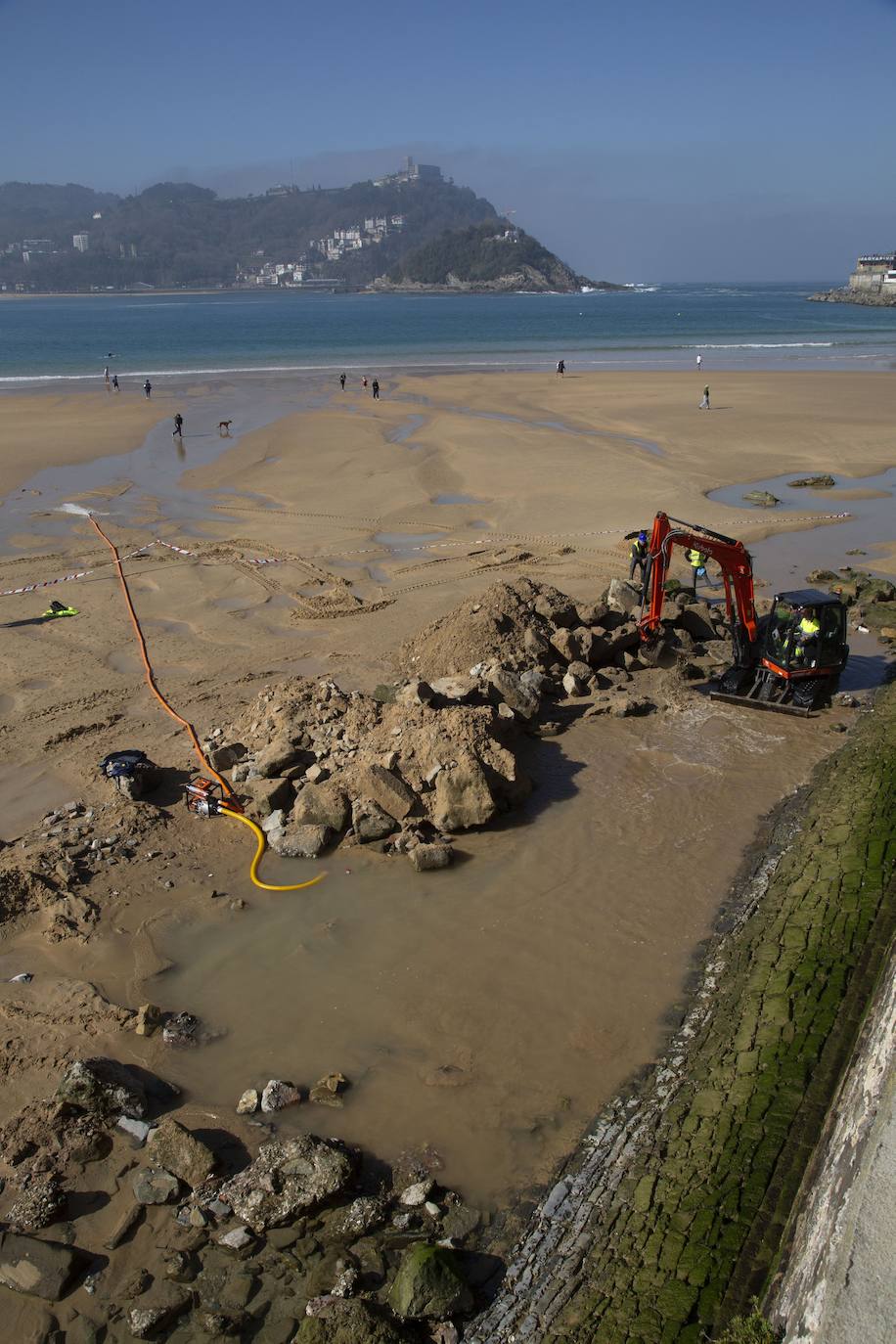 La playa de La Concha se ha convertido en la bajamar de este lunes por la mañana en un inesperado yacimiento arqueológico. La presencia de una pequeña excavadora retirando piedras y escombros junto al muro de costa a la altura del edificio consistorial ha suscitado una gran expectación entre los numerosos viandantes y turistas que han provechado el buen tiempo para dar un paseo. La máquina profundizaba en la arena en busca de los restos de un barco hundido del que hay constancia documental desde el siglo XVIII. 