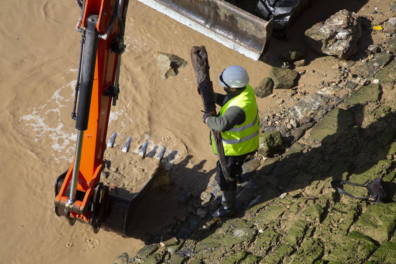La playa de La Concha se ha convertido en la bajamar de este lunes por la mañana en un inesperado yacimiento arqueológico. La presencia de una pequeña excavadora retirando piedras y escombros junto al muro de costa a la altura del edificio consistorial ha suscitado una gran expectación entre los numerosos viandantes y turistas que han provechado el buen tiempo para dar un paseo. La máquina profundizaba en la arena en busca de los restos de un barco hundido del que hay constancia documental desde el siglo XVIII. 
