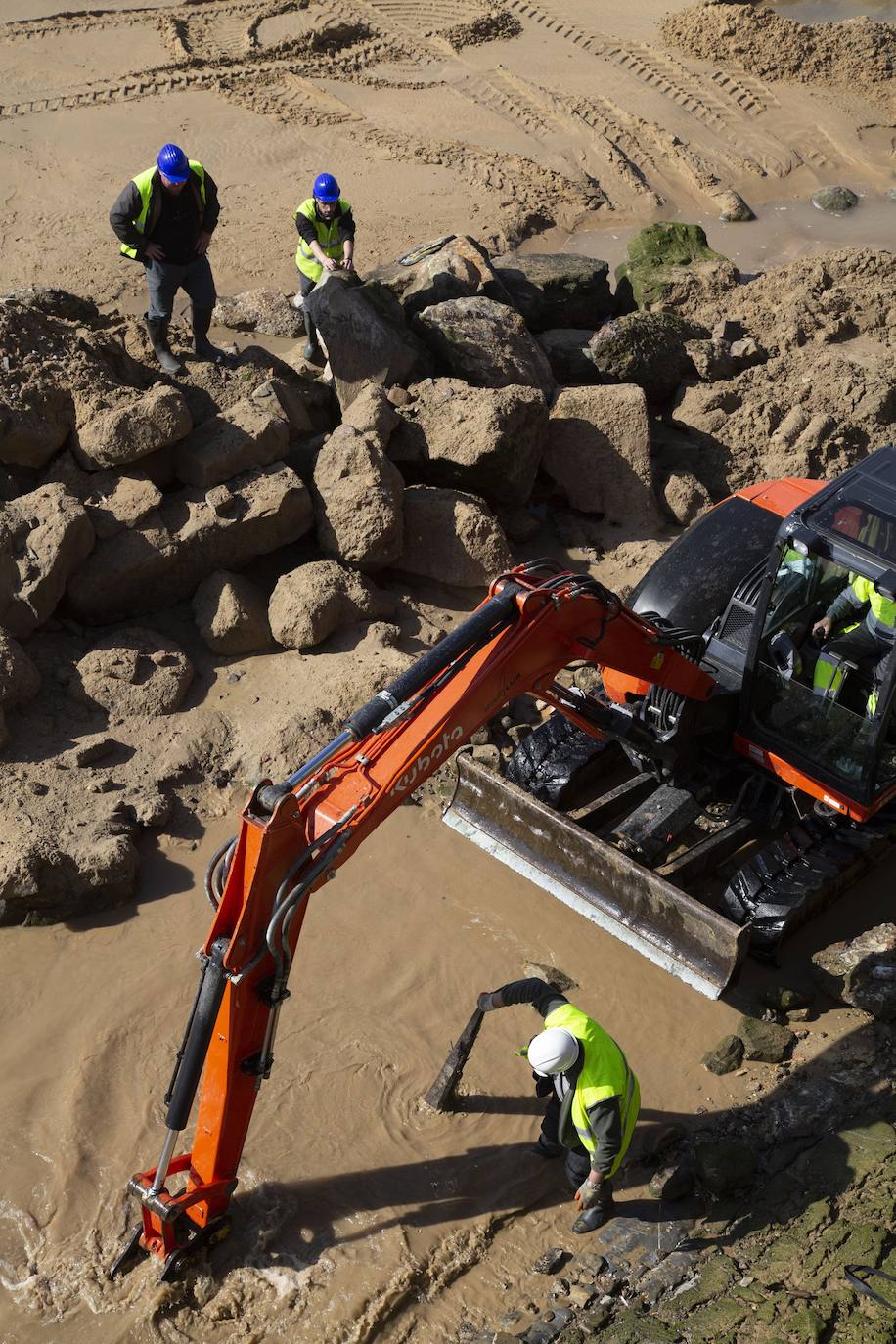 La playa de La Concha se ha convertido en la bajamar de este lunes por la mañana en un inesperado yacimiento arqueológico. La presencia de una pequeña excavadora retirando piedras y escombros junto al muro de costa a la altura del edificio consistorial ha suscitado una gran expectación entre los numerosos viandantes y turistas que han provechado el buen tiempo para dar un paseo. La máquina profundizaba en la arena en busca de los restos de un barco hundido del que hay constancia documental desde el siglo XVIII. 