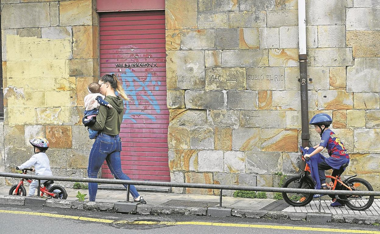 Una mujer camina con un bebé en brazos por una calle de Zumarraga, con otros dos niños montando en bicicleta.