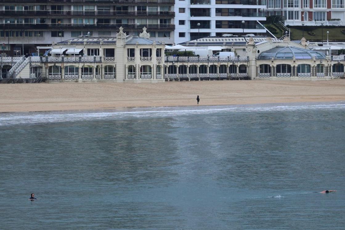 Dos nadadores surcan las aguas de La Concha mientras otra persona observa la bahía desde la orilla.