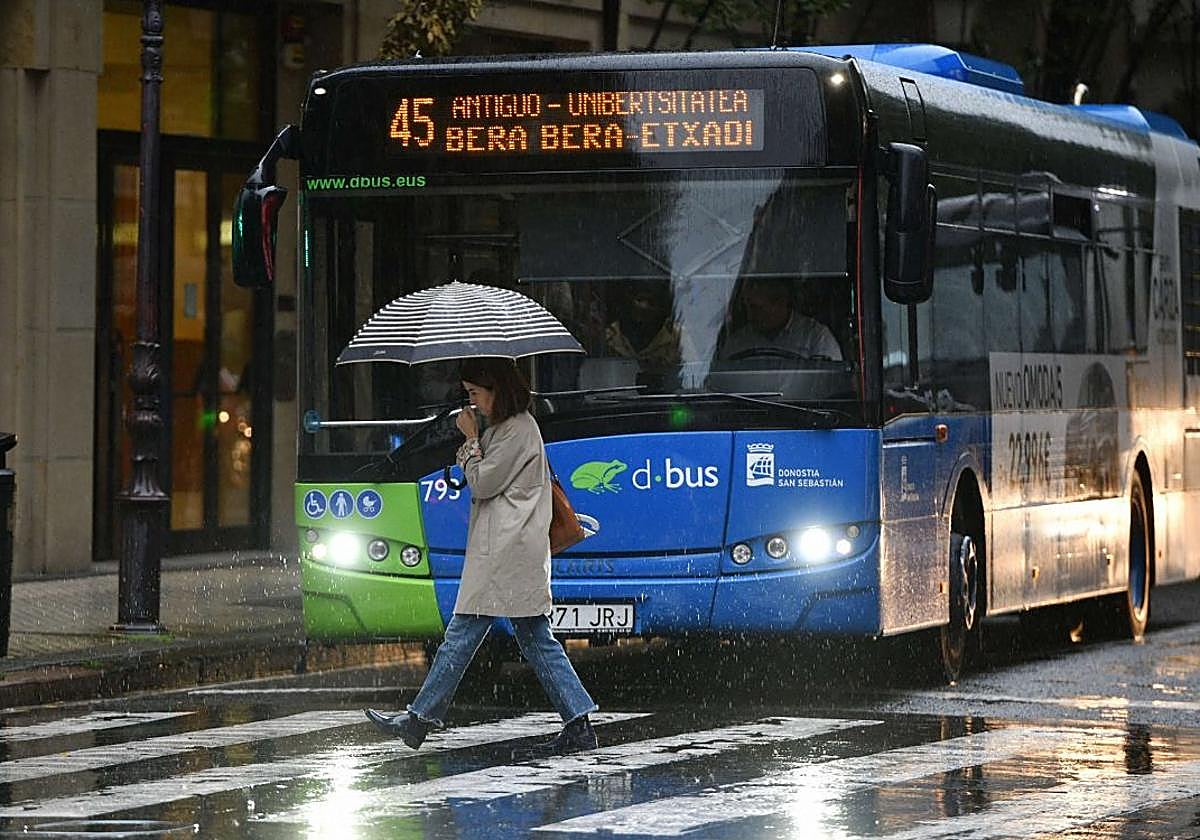 Una mujer cruza un paso de peatones frente a un autobús de Dbus mientras se refugia bajo un paraguas en un día de lluvia en San Sebastián.