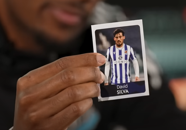 Alex Isak sujetando el cromo de David Silva de txuri-urdin.