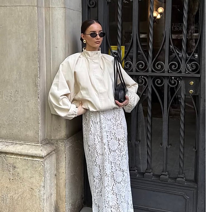 Cazadora crema de cuello alto y mangas voluminosas sobre falda midi de encaje floral y bolso mini negro.