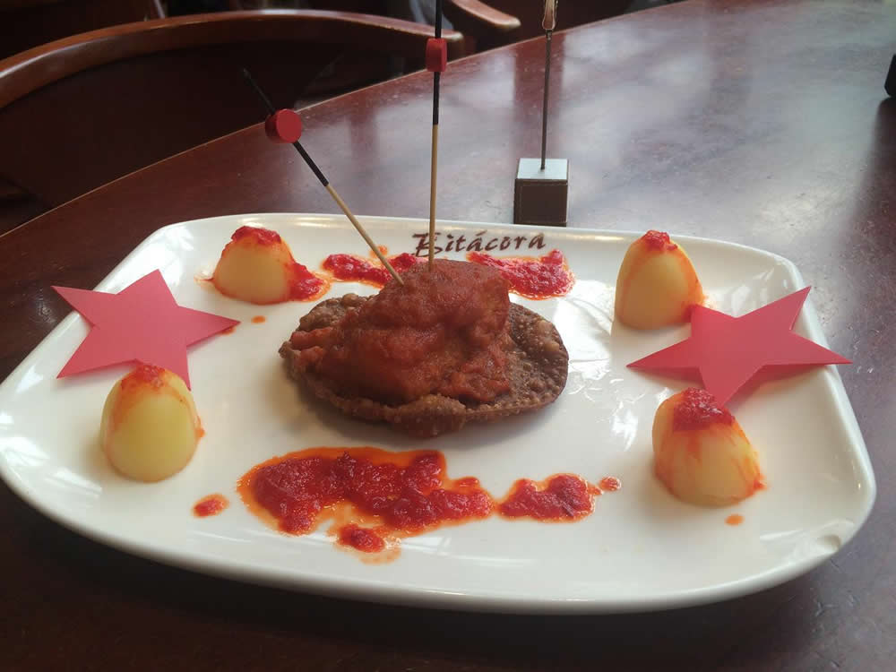 Bocadito de bacalao (Bitácora). Bacalao en base de oblea crujiente, tomate natural, patatas al vapor y acompañado de una salsa de pimiento agridulce.