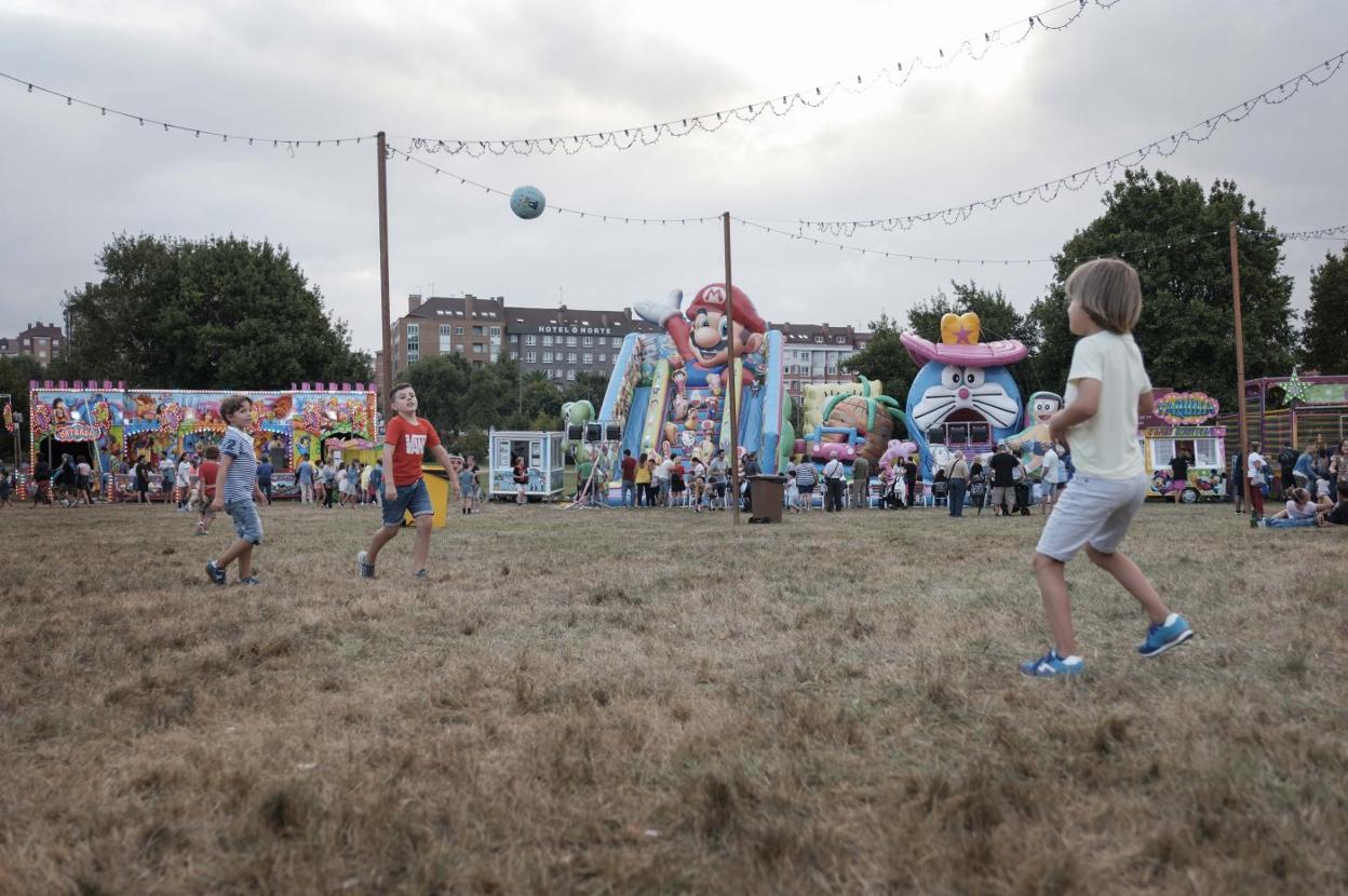 Niños juegan al balón en el prau de la fiesta de Contrueces frente a los tiovivos. 