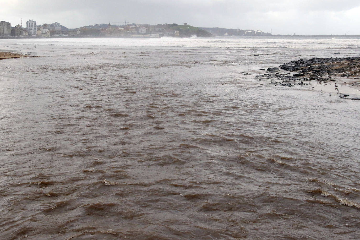 El temporal que azota Asturias ha ocasionado que la bahía de San Lorenzo apareciera este viernes con agua de color marrón, procedente del río Piles y del arrastre de las aguas de escorrentía, lo que se unió a una mar muy revuelta