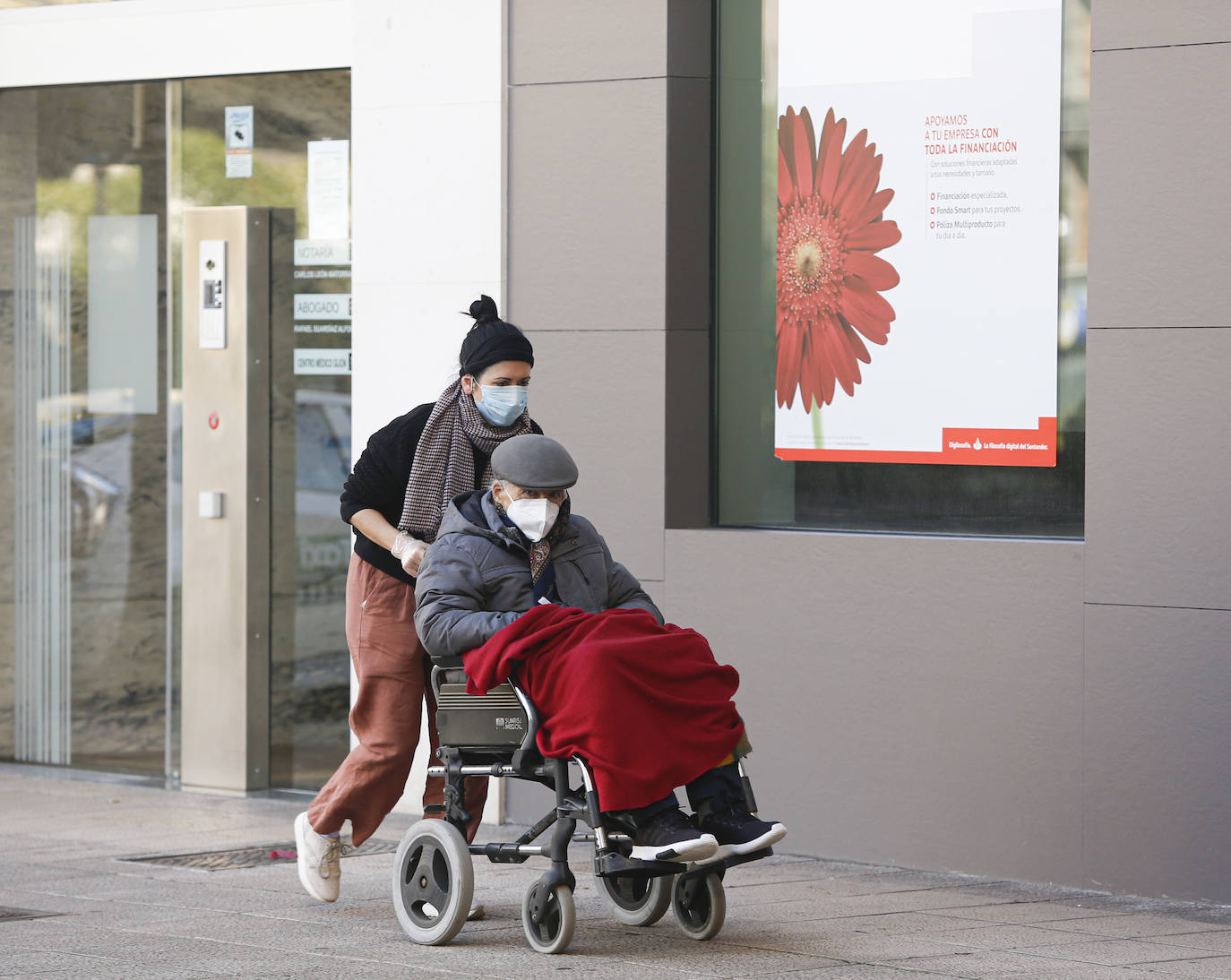07 de abril. Gijón | Una mujer empuja la silla de ruedas de un anciano, ambos con mascarillas. 