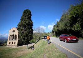 Un hombre pasea por el entorno de Santa María del Naranco junto a la carretera, por la que circula un vehículo.