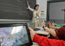 Una profesora guía una clase desde la pizarra clásica mientras sus alumnos siguen sus indicaciones en tablets.