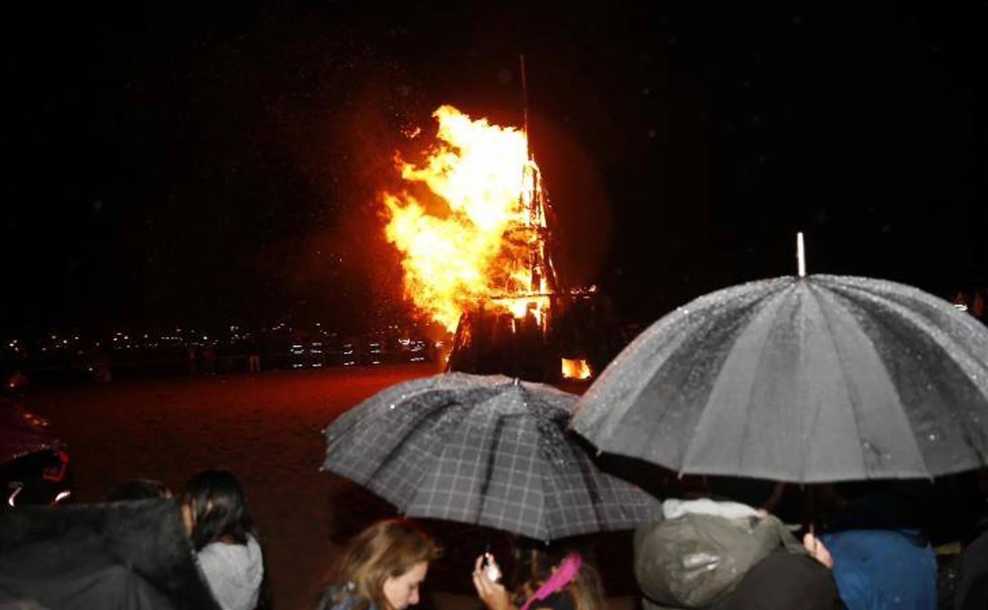 Los vecinos, bajo el paraguas, contemplan el fuego en la playa de Poniente, en Gijón.
