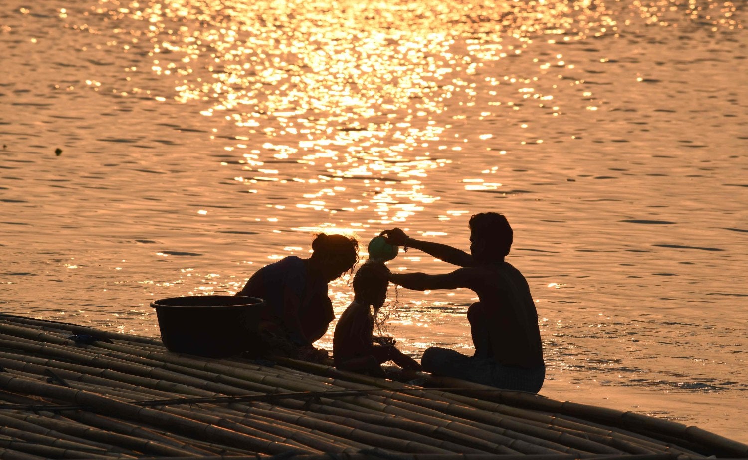 Un hombre indio baña a su hijo al atardecer mientras se sienta sobre cañas de bambú en venta a lo largo del río Brahmaputra, en Guwahati