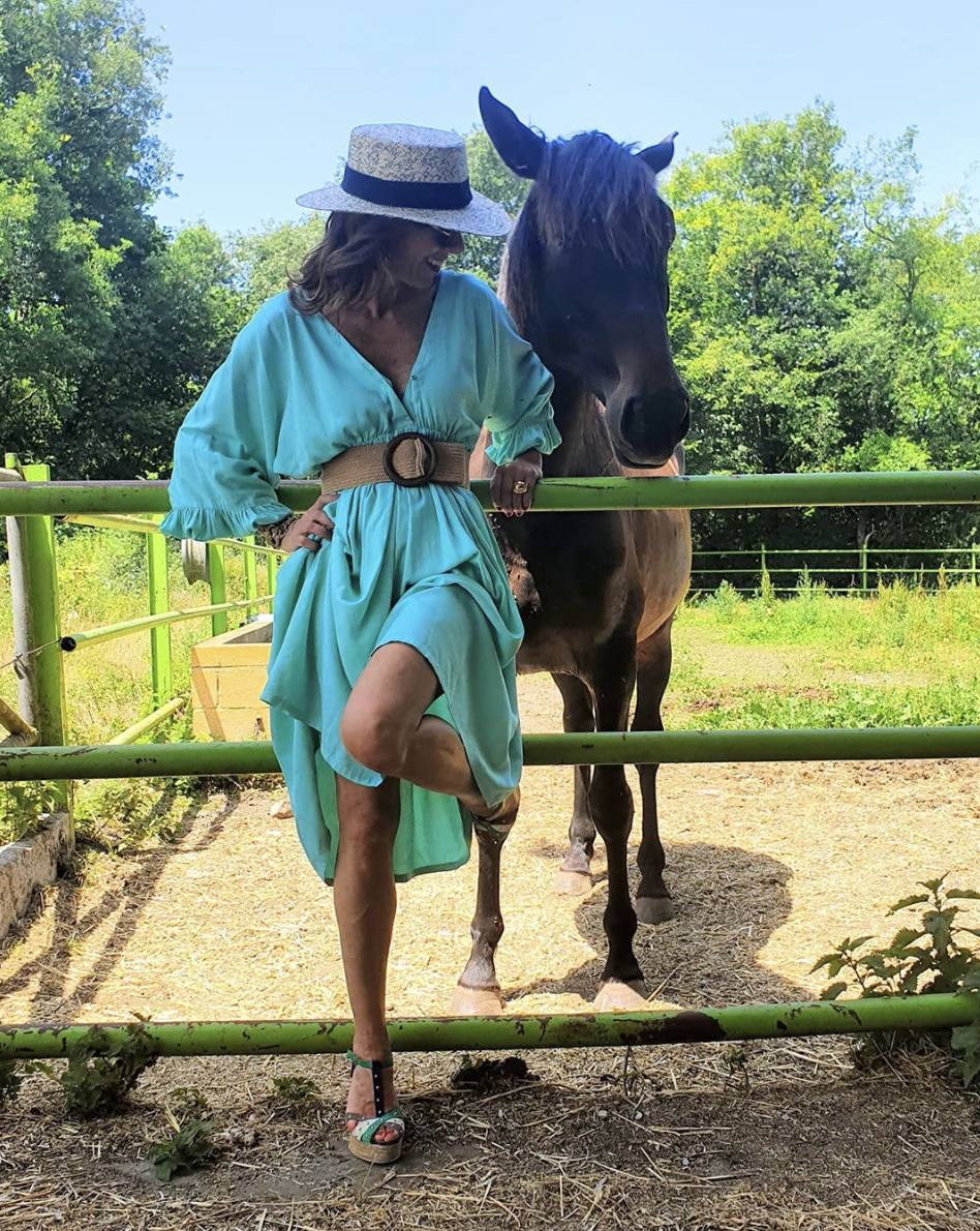 Se fotografía así de estupenda en el campo en compañía de un caballo. Su look está compuesto por un vestido azul cielo ceñido con un cintuón de rafia. Lo combina con unas cuñas de esparto y un sombrero Panamá.