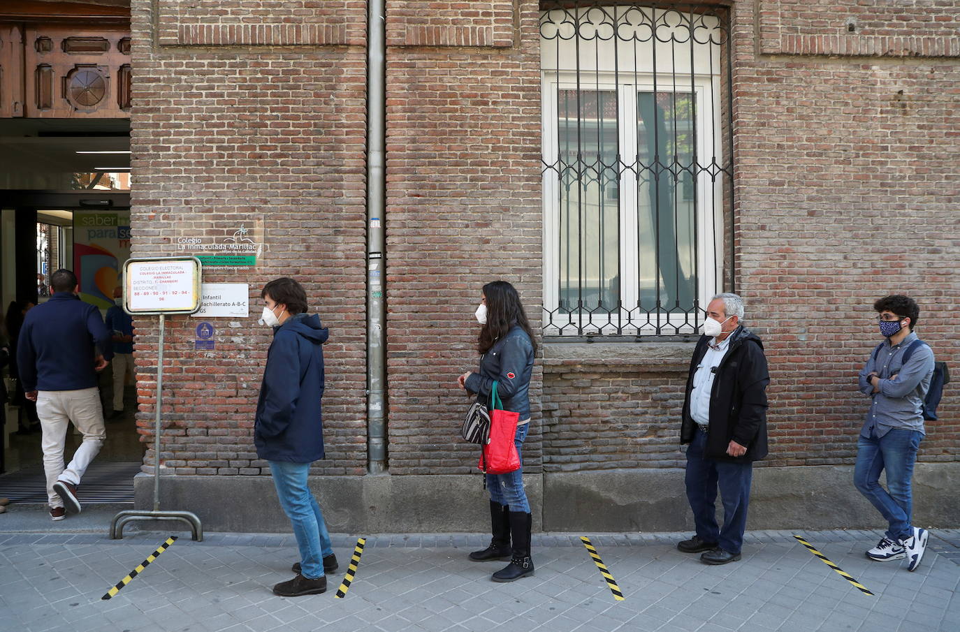 Los votantes, con máscaras protectoras y manteniendo la distancia social, hacen cola fuera de un colegio electoral durante las elecciones a la Comunidad de Madrid.