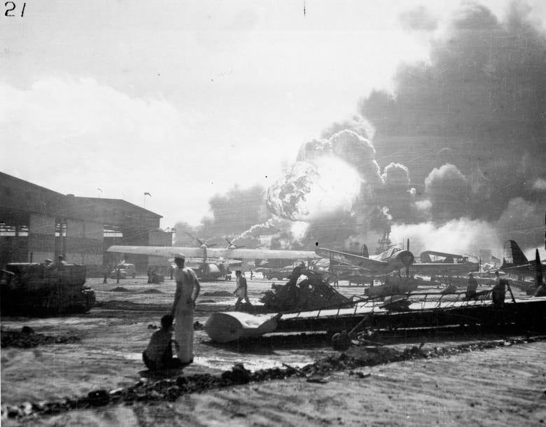 Los marineros se paran en medio de aviones destrozados en la base de hidroaviones de la isla Ford, viendo el destructor USS Shaw explotar en el fondo del centro. Archivos Nacionales / Marina de los EE. UU.