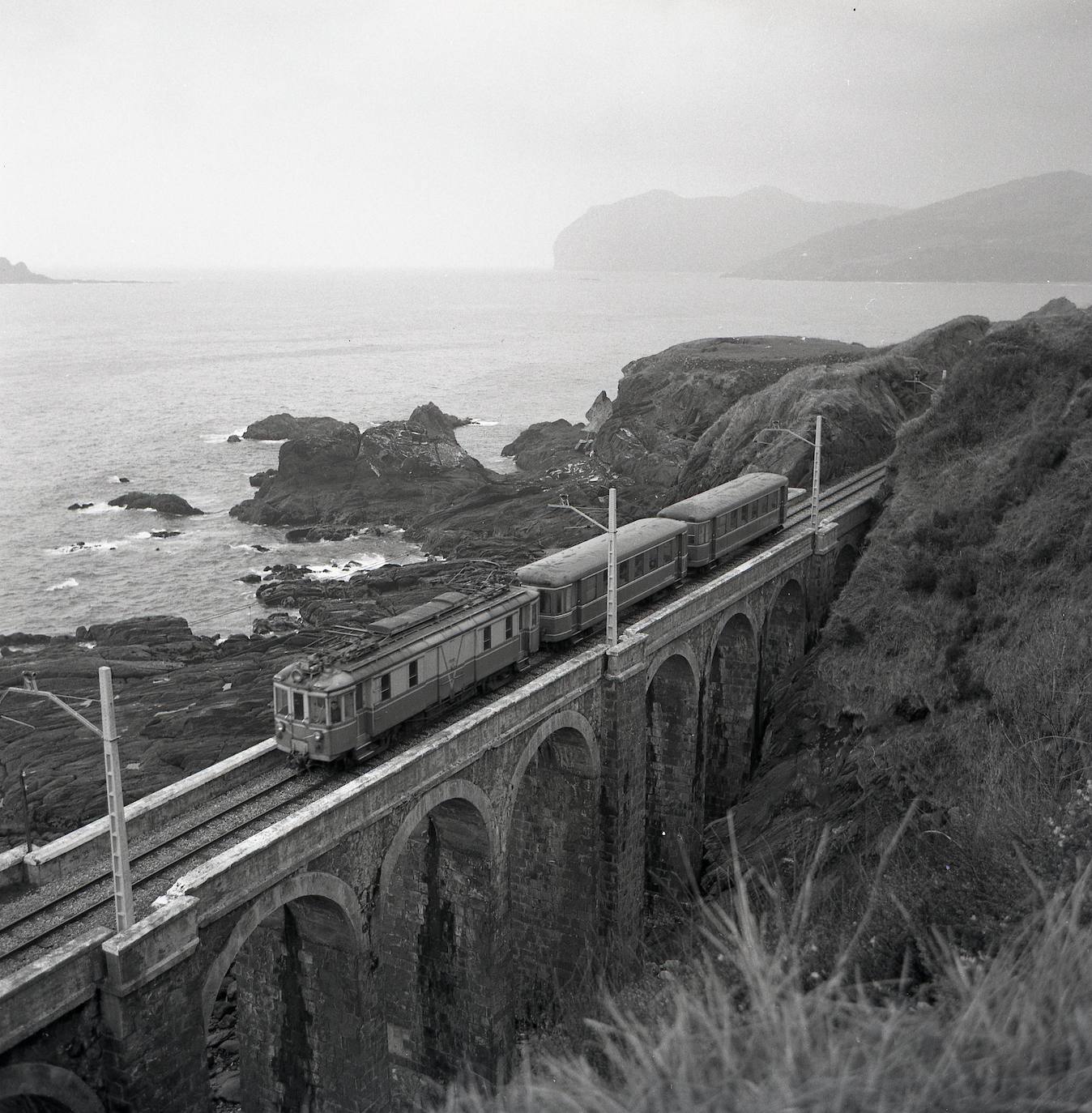 Imagen principal - Un tren pasa por el viaducto, con la isla de Izaro arriba a la izquierda y con el cabo de Ogoño, a la derecha. Abajo, el día de la inauguración de la prolongación del tren desde Sukarrieta, un acto al que asistió Franco. A la derecha, otra estampa del tren entrando en Bermeo. 