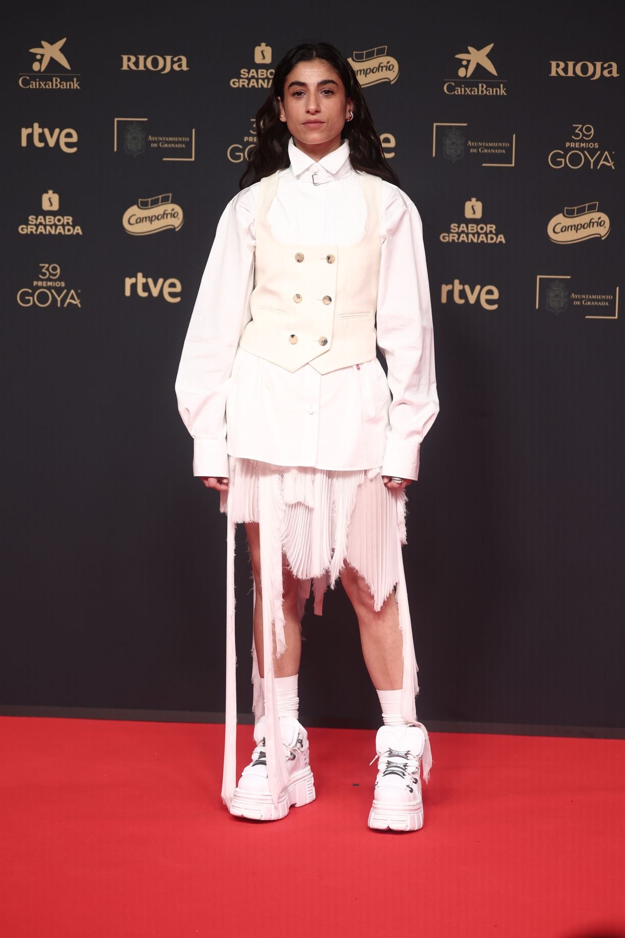 La actriz Carolina Yuste ha presumido de estilo propio con un look en blanco con el sello de Alexander McQueen. Combina una camisa 'oversize' con un chaleco de estilo sastre y una falda plisada desestructurada. La artista ha marcado la diferencia eligiendo unas botas de suela track con aplicaciones metálicas sobre las que asoman unos calcetines a tono. En cuanto a las joyas, ha optado por piezas de Suot que aportan un toque plateado al 'outfit'.