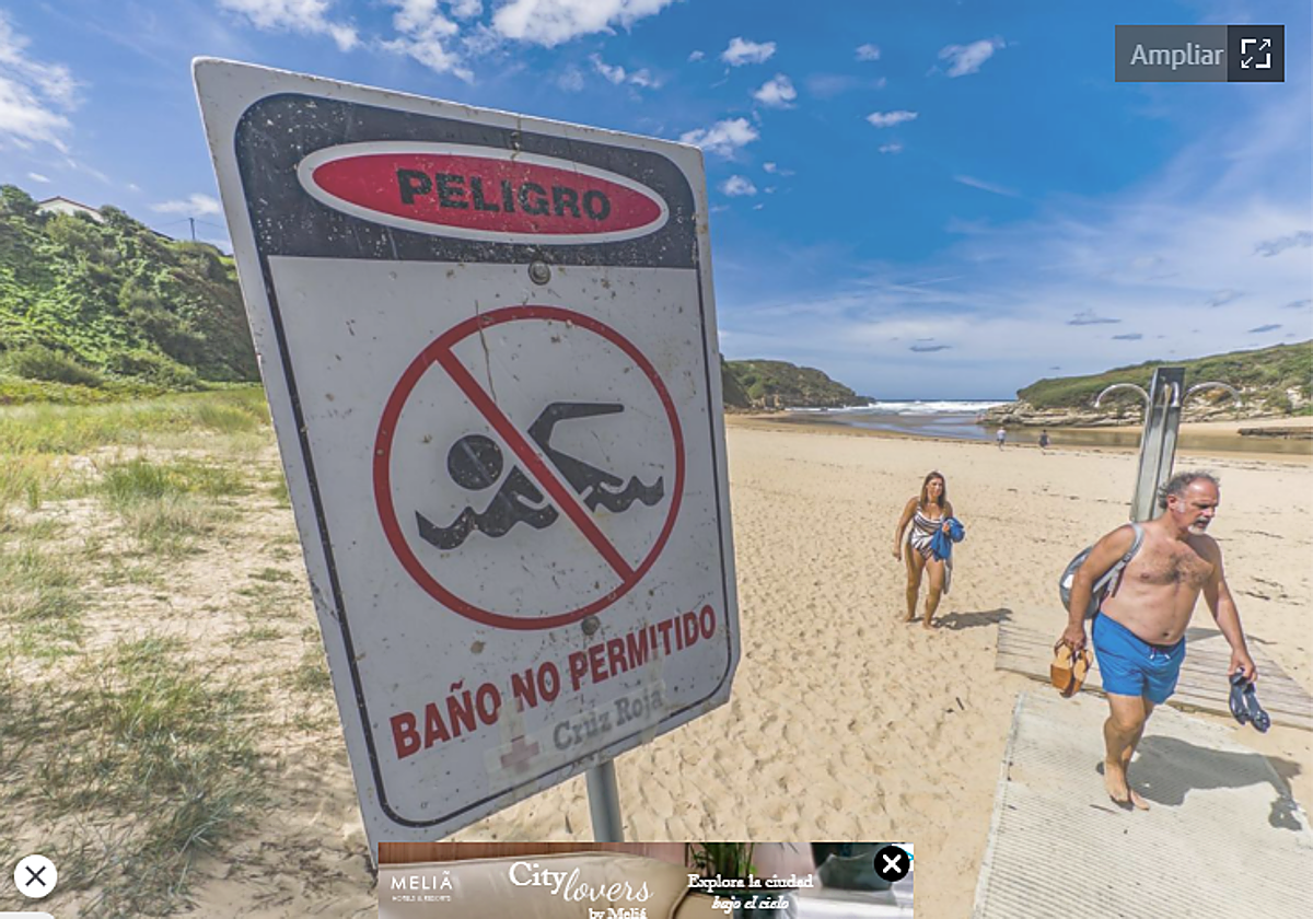 Dos personas abandonan la playa de Galizano, donde un cartel indica la prohibición de bañarse por contaminación.