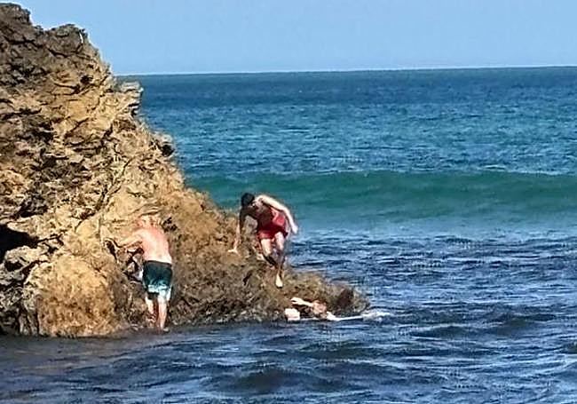 El hombre se agarra a la roca, mientras en el agua el ertzaintza sujeta a la mujer exhausta y el hombre del bañador rojo se acerca para ayudarlos.