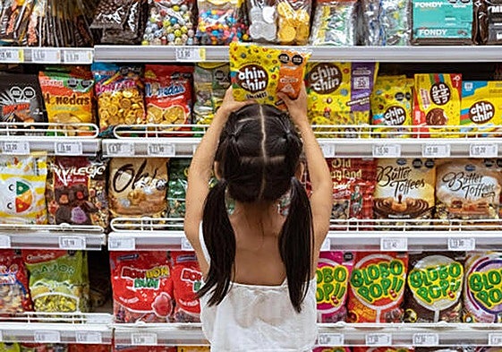 Una niña coge una bolsa de chucherías en una estantería con aperitivos dulces y salados de un supermercado.