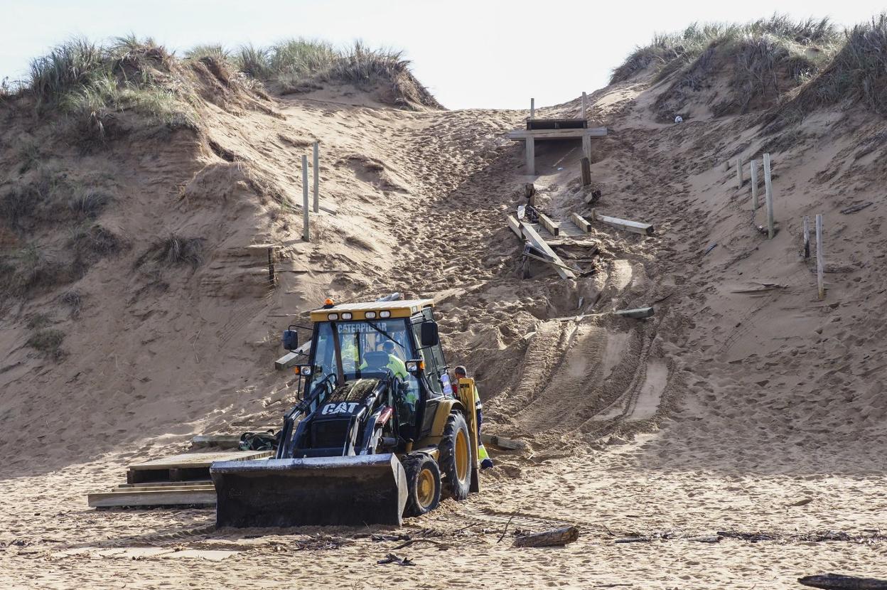 Una máquina retira las tablas de madera de la pasarela que se había derrumbado en el sistema dunar de la playa de Valdearenas. 