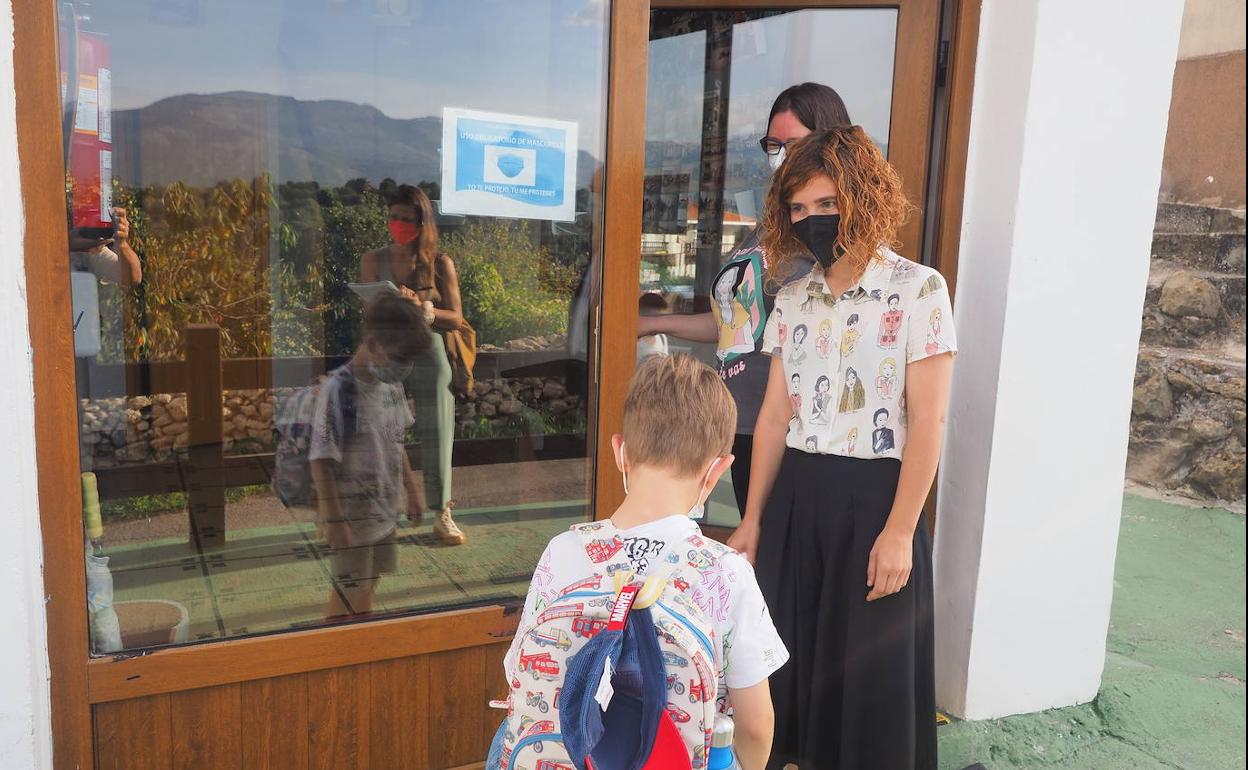 Una profesora con mascarilla recibe a un alumno en la puerta del centro educativo. 