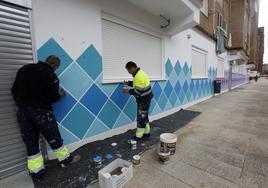 Dos trabajadores pintan un mural artístico en un local municipal de Nueva Ciudad.