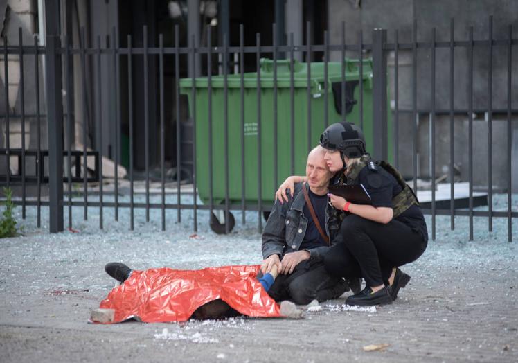 Imagen principal - 1. Un padre sujeta la mano de su hijo de 13 años, muerto en una parada de autobús de la ciudad de Jersón por un bombardeo ruso en julio de 2022. A su lado, una policía ucraniana le consuela. | 2. Funeral colectivo por veinticinco soldados ucranianos muertos en el frente este de la guerra contra Rusia. | 3. Ciudadanos tejen redes miméticas (de camuflaje) para el ejército ucraniano en una calle en Leópolis.