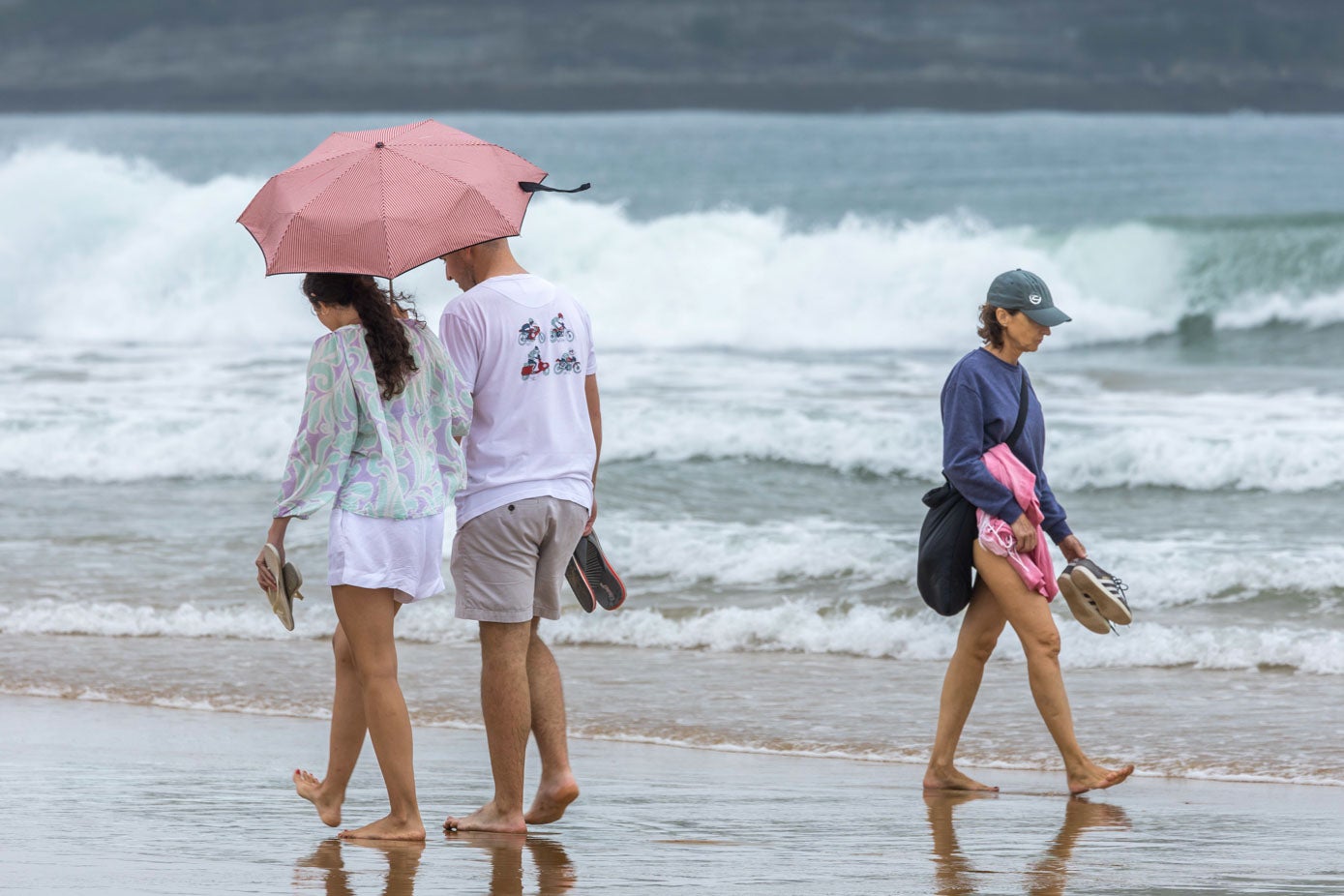 Dos jóvenes turistas se protegen de la lluvia con un paraguas por la orilla de la playa de El Sardinero. 