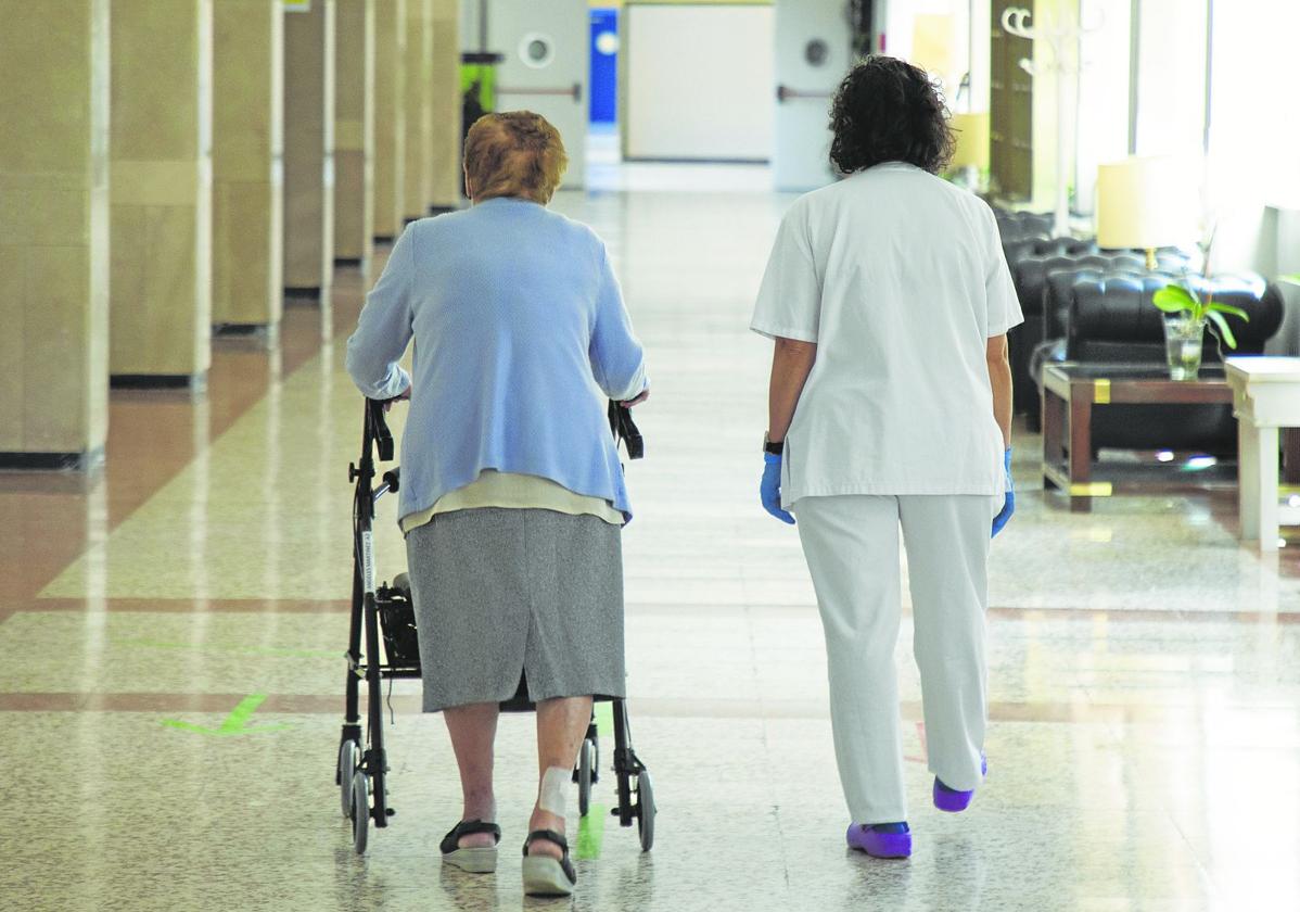 Una mujer pasea por el interior de una residencia de ancianos acompañada por una cuidadora.