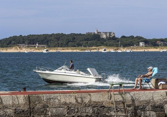 Un hombre toma el sol en el puerto de Pedreña mientras una motora abandona la localidad.