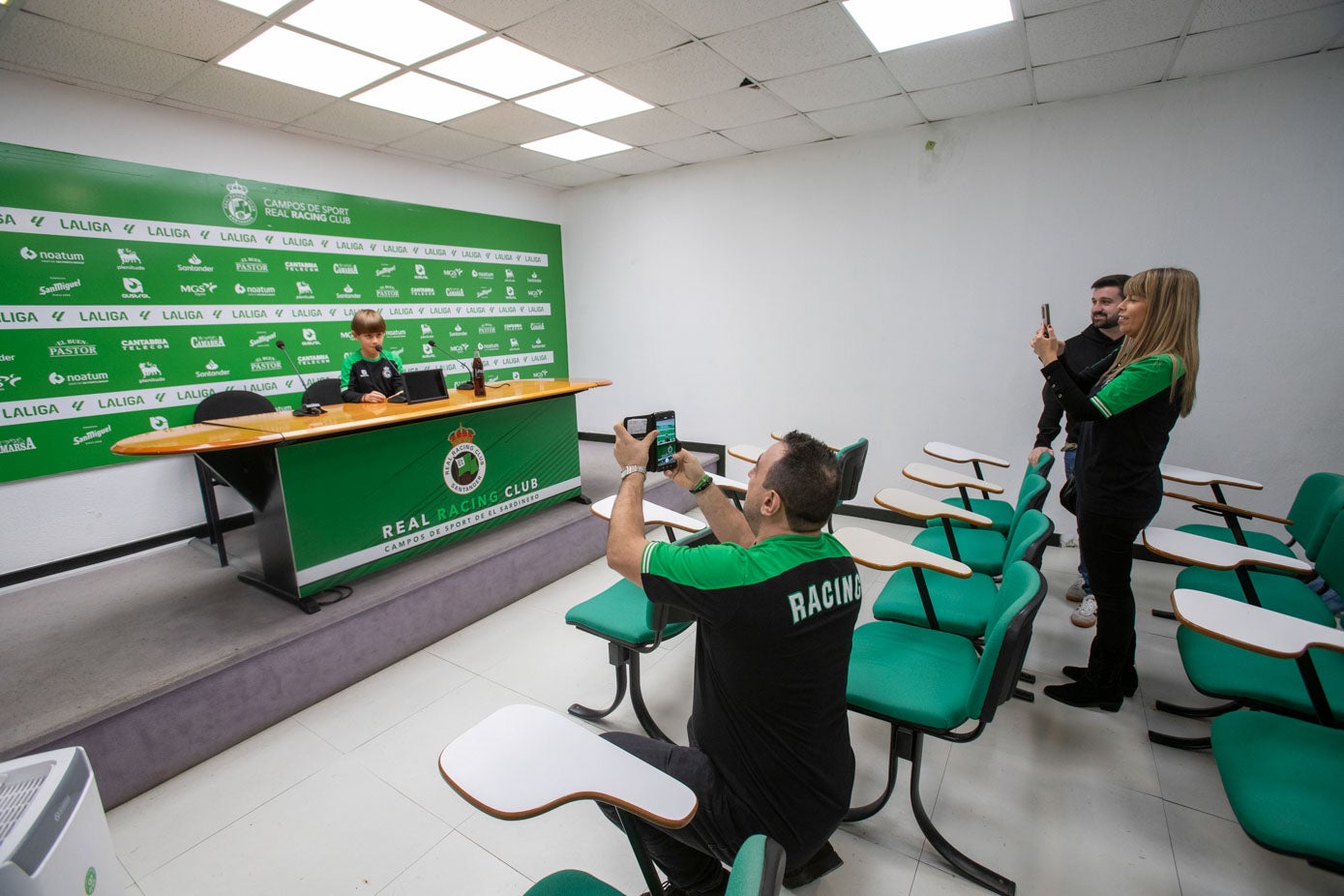 Roberto Velo, sentado en la sala de prensa, simula ofrecer declaraciones mientras sus padres le toman fotografías. 