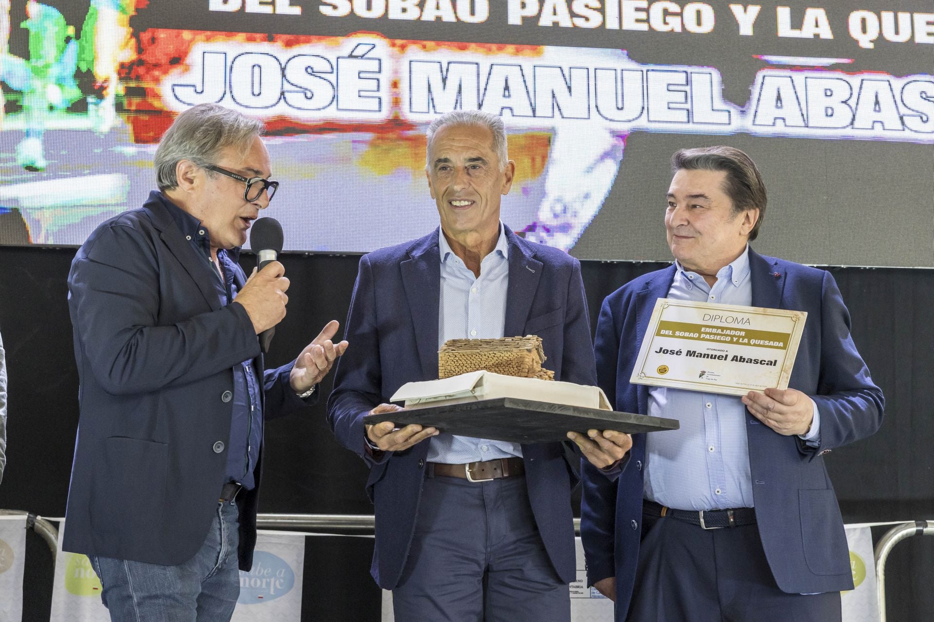 El alcalde de Vega de Pas, Juan Carlos García (izquierda), y el presidente de la IGP de Sobao Pasiego, Adolfo Gómez (derecha), entregan a José Manuel Abascal la escultura de una cabaña pasiega sobre la base de un sobao, como símbolo de los Valles Pasiegos.