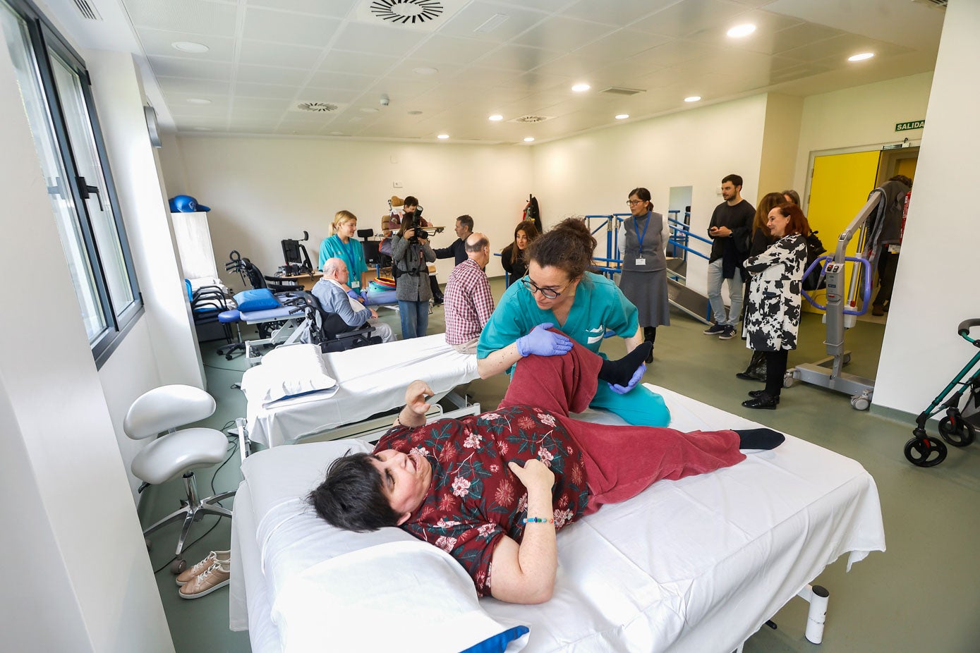 Una trabajadora realiza estiramientos con una de las usuarias del CAD, en primer término, con trabajadores y autoridades al fondo, durante la visita de la Consejería.