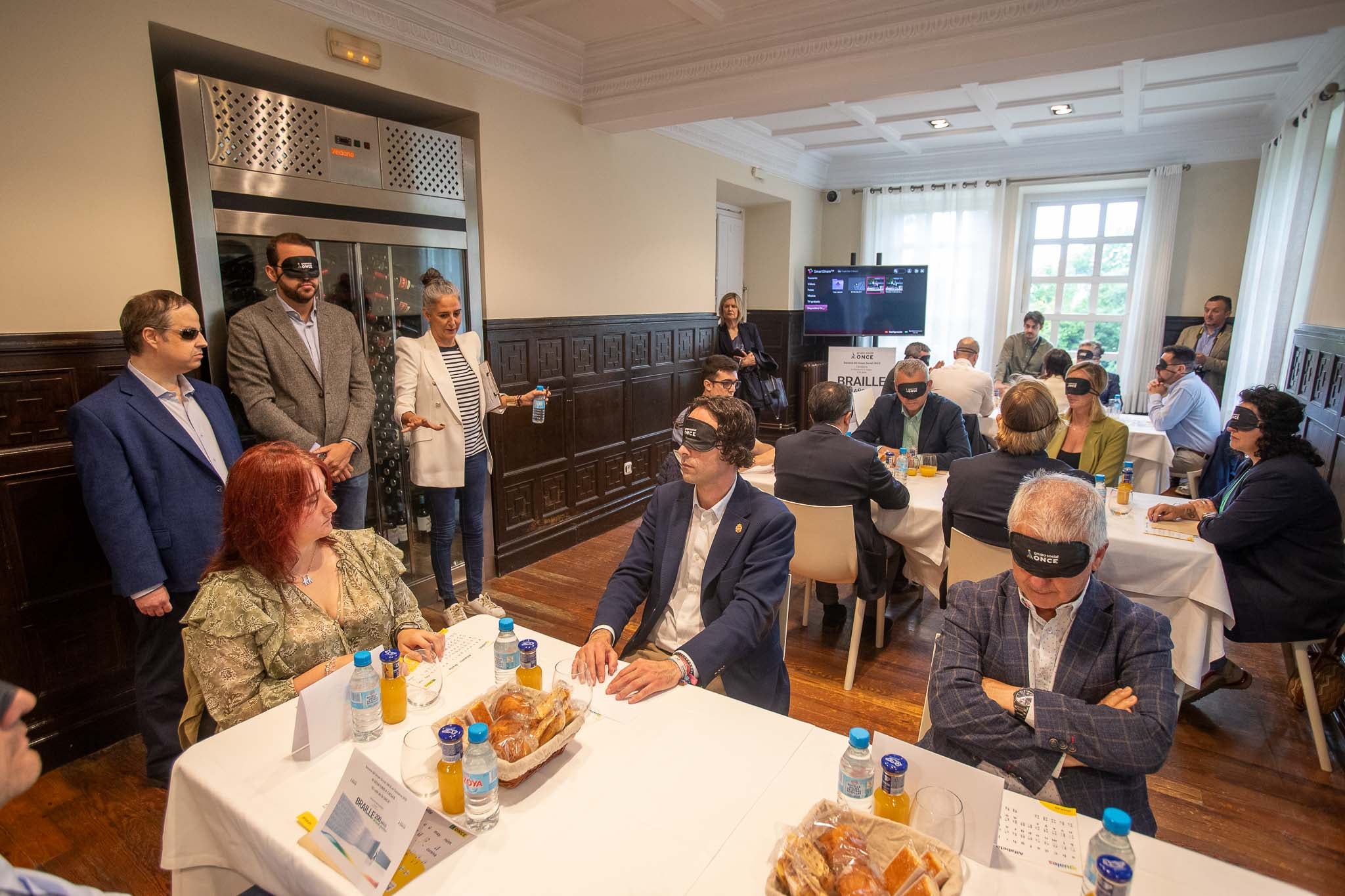 La veintena de alcaldes entraron al desayuno con los ojos tapados. En primer plano Javier Fernández Soberón (Astillero) junto a Constantino Fernández (Villaescusa). A la izquierda, la afiliada de la ONCE Zoe Laso. Bajo la mesa está su perro guía Gaby. De pie, el presidente de la ONCE en Cantabria, Sergio Olavarría; el presidente de los hosteleros cántabros, Eduardo Lamadrid; y la periodista Leticia Mena, que presentó el evento.