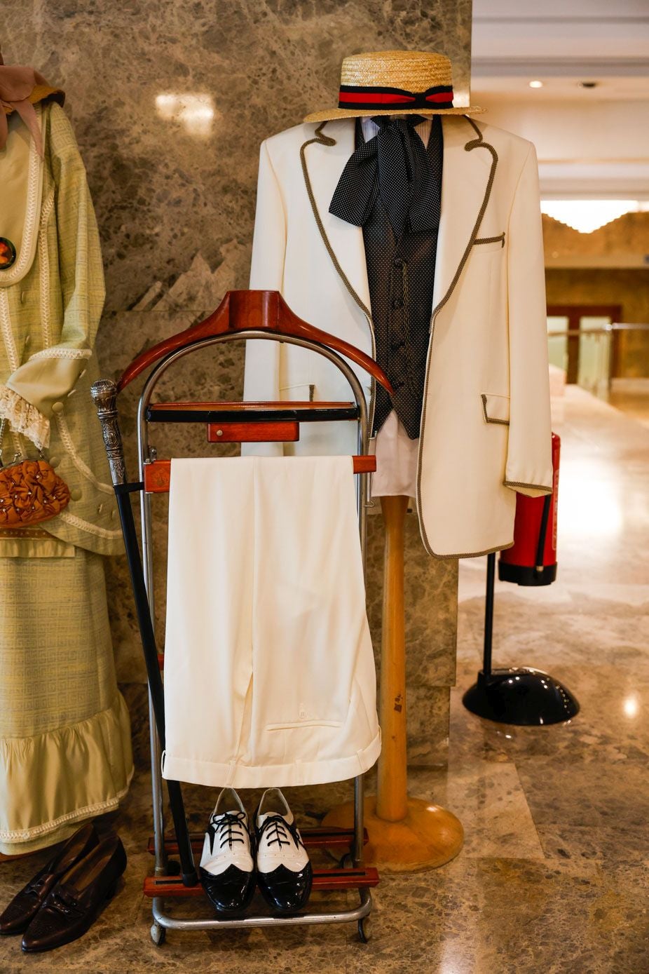 Traje de caballero blanco con chaqueta ribeteada con pasamanería, chaleco, camisa y lazada al cuello. Se corona el conjunto con un sombrero de esparto adornado con una cinta negra y roja.